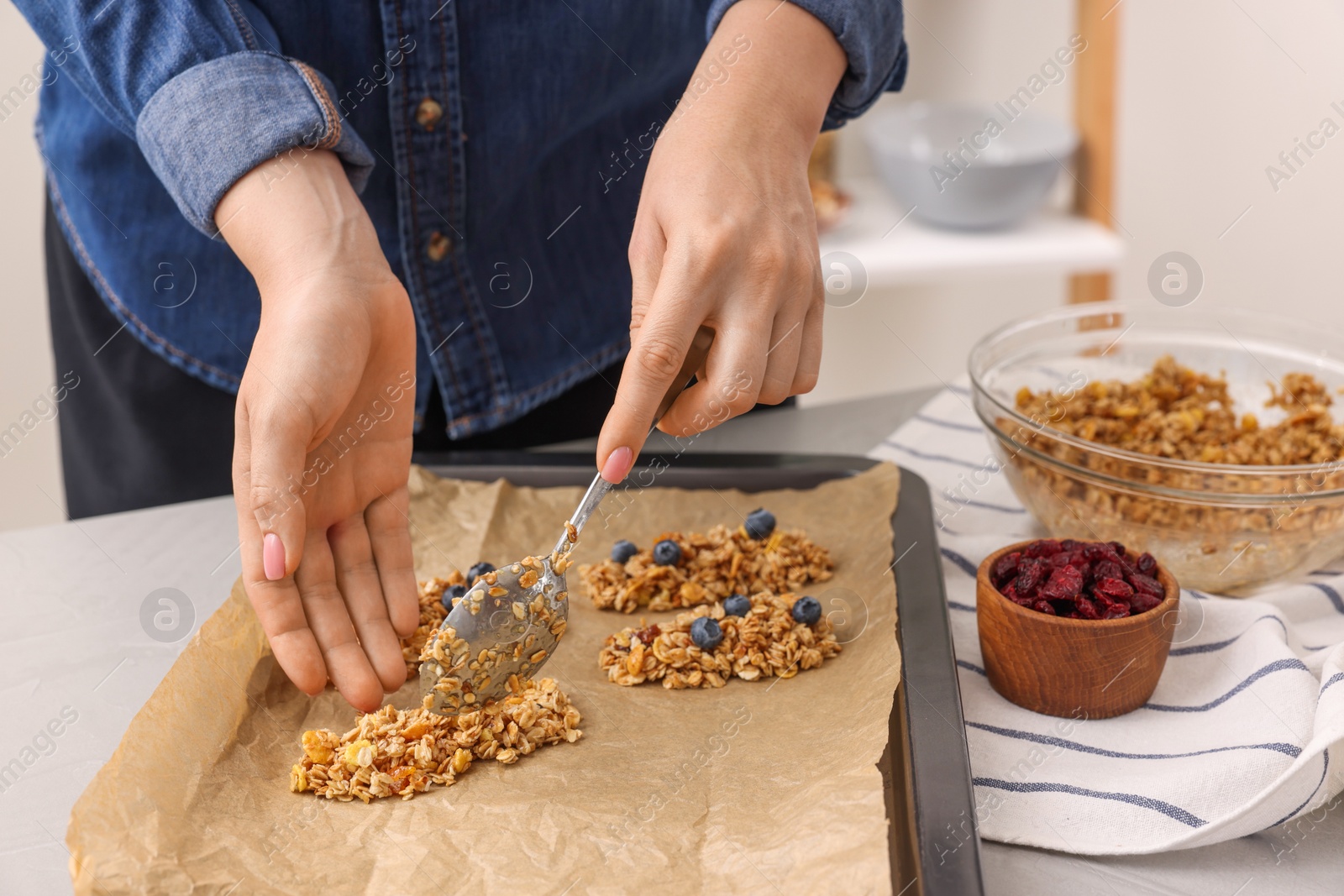 Photo of Making granola bars. Woman putting mixture of oat flakes, dry fruits and other ingredients onto baking tray at table in kitchen, closeup