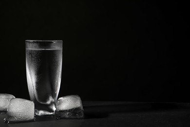 Photo of Vodka in shot glass with ice on table against black background. Space for text