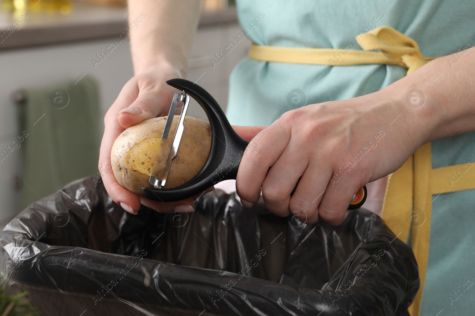 Photo of Woman peeling fresh potato above garbage bin indoors, closeup