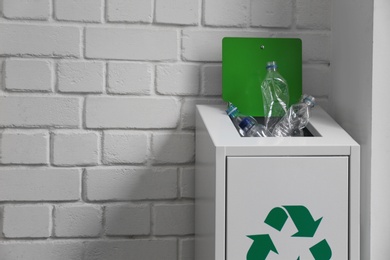 Many used plastic bottles in metal bin near white brick wall, space for text. Recycling problem