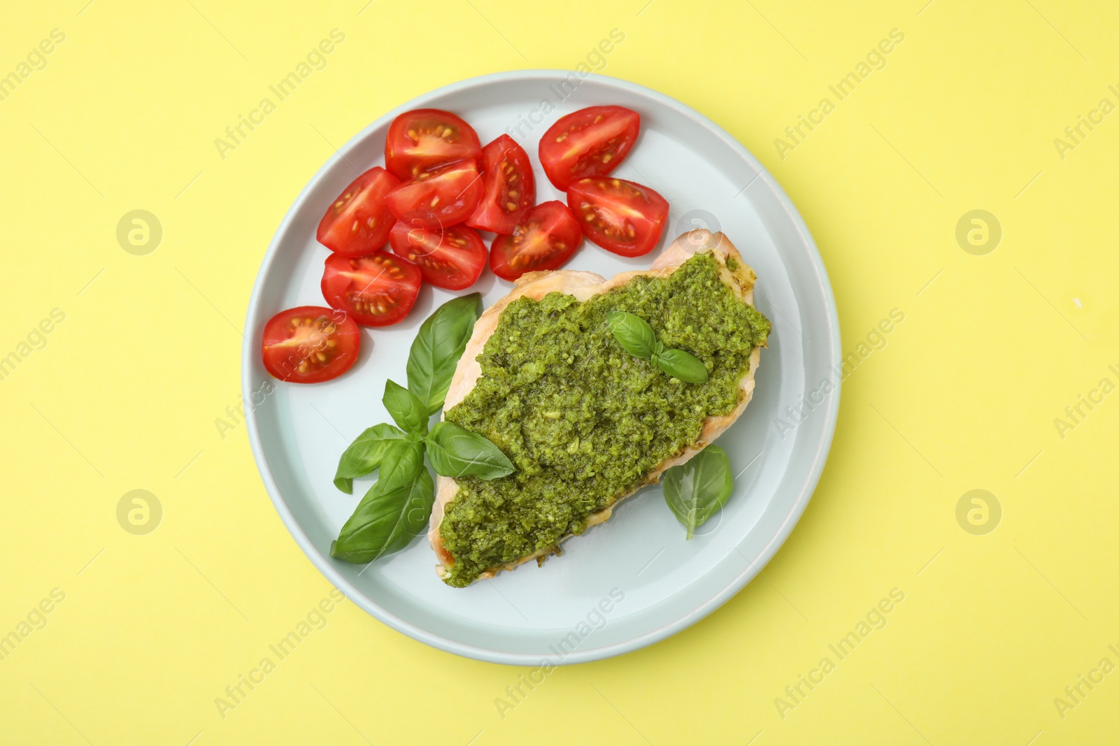 Photo of Delicious chicken breast with pesto sauce, tomatoes and basil on yellow table, top view