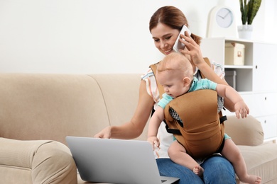 Photo of Woman with her son in baby carrier using laptop and talking on phone at home. Space for text