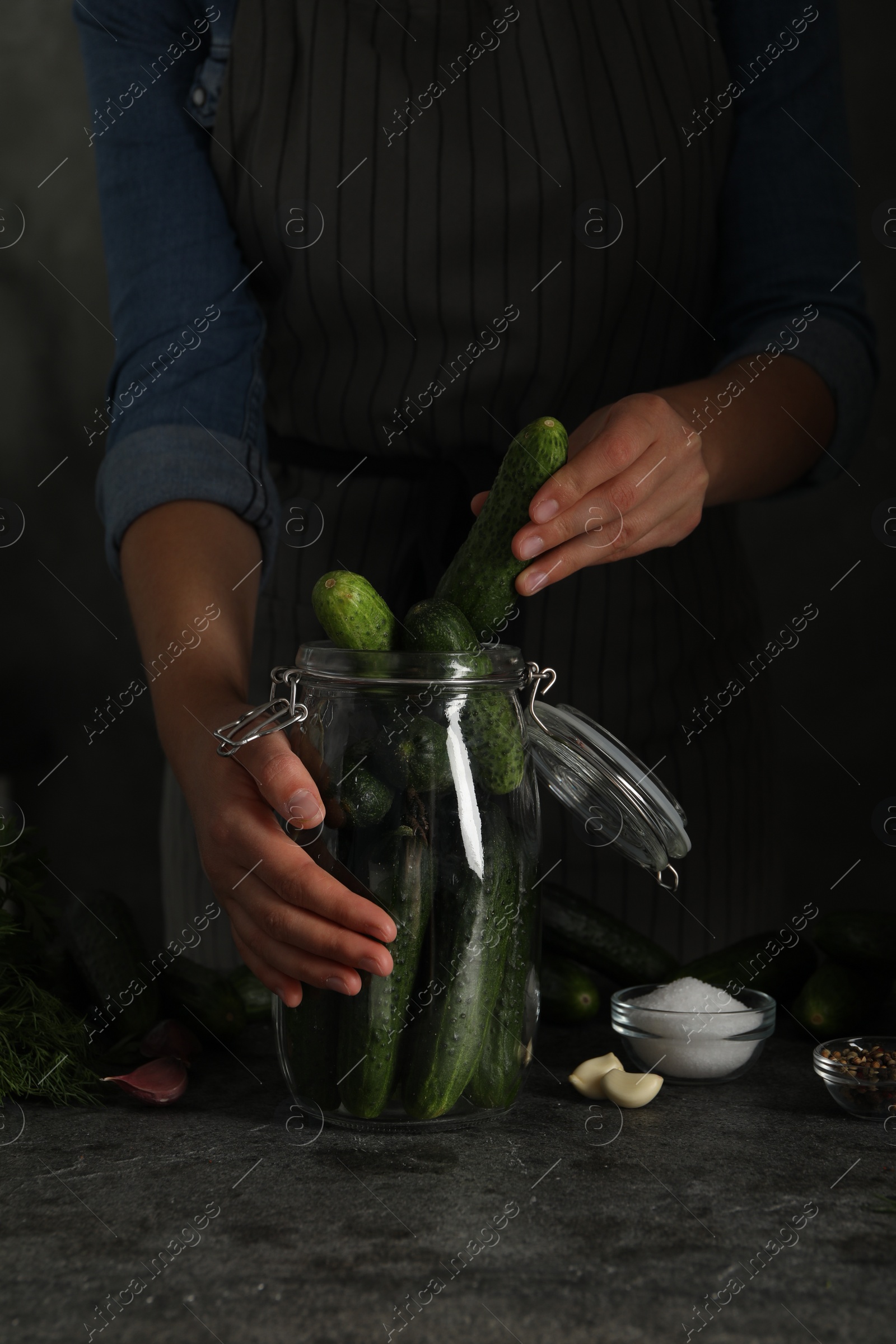 Photo of Woman putting cucumber into glass jar at black kitchen table, closeup. Pickling vegetables