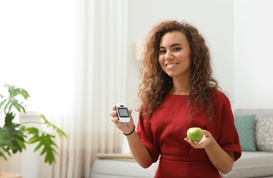 Photo of Young African-American woman holding digital glucometer and apple at home. Diabetes diet