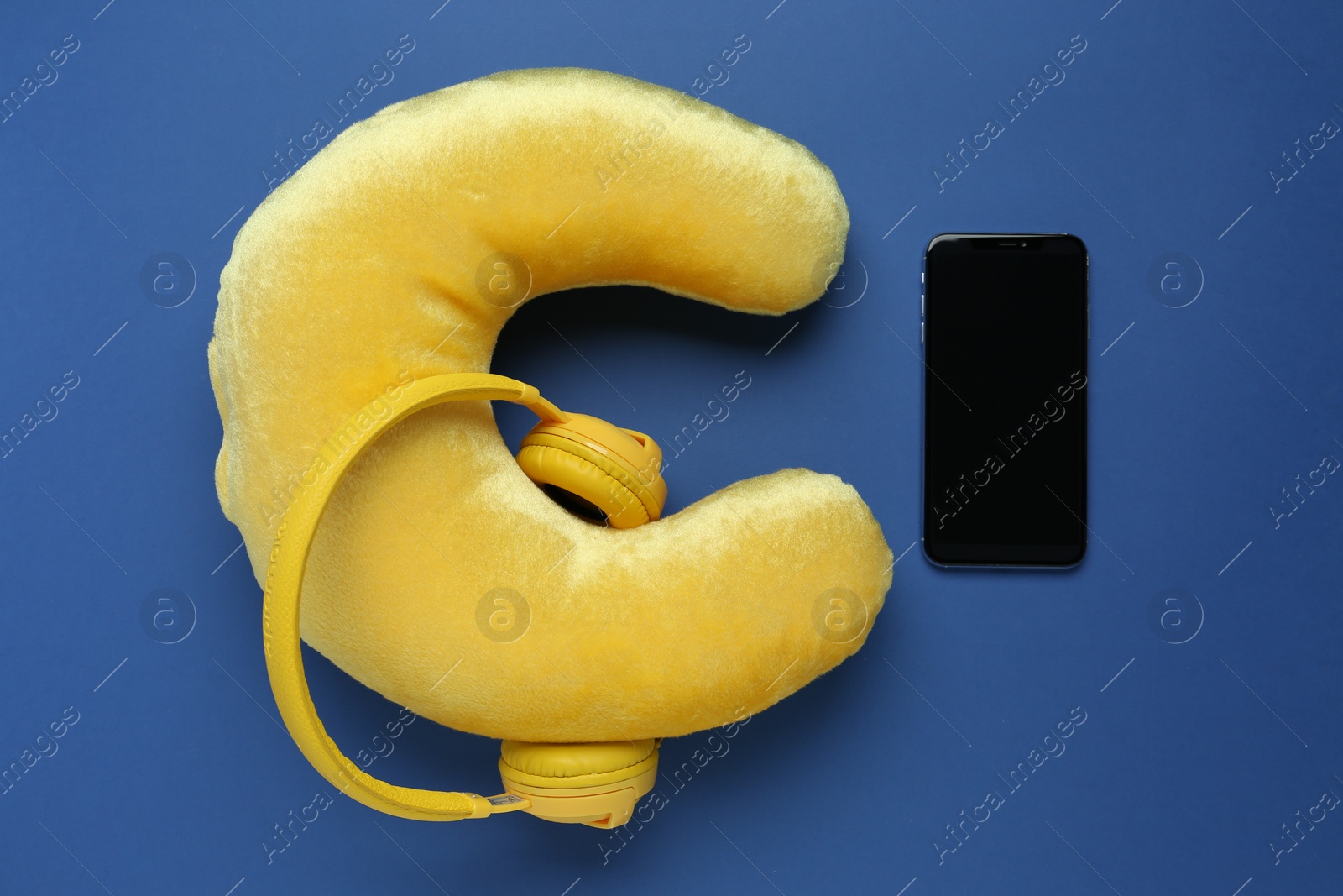 Photo of Yellow travel pillow, smartphone and headphones on blue background, flat lay