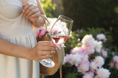 Photo of Woman with glass of rose wine and straw bag in peony garden, closeup