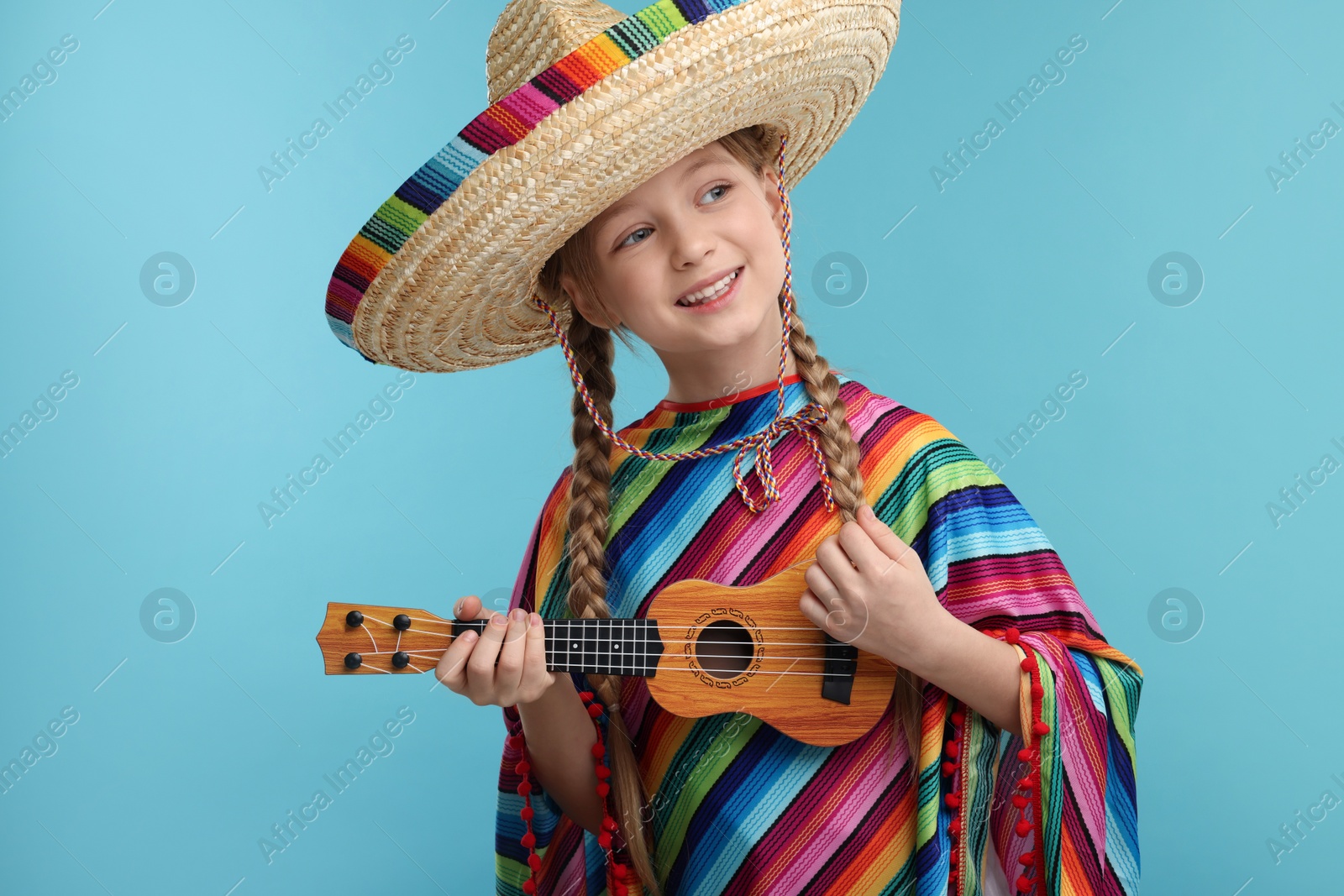Photo of Cute girl in Mexican sombrero hat and poncho playing ukulele on light blue background