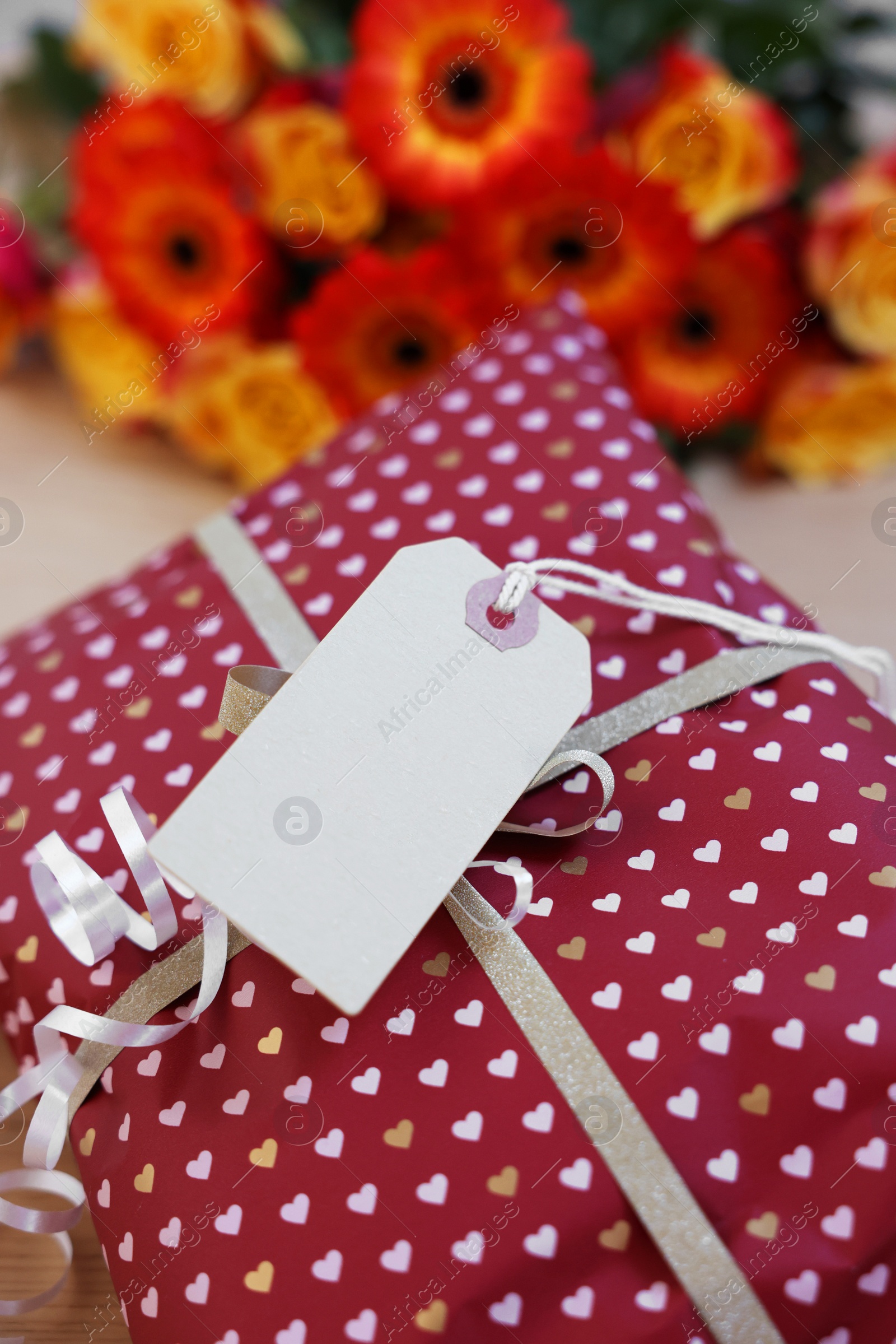 Photo of Parcel wrapped in heart patterned paper with cardboard tag and beautiful flowers on table, closeup. Space for text