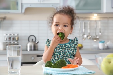 Photo of Cute African-American girl eating vegetables at table in kitchen