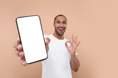 Photo of Young man showing smartphone in hand and OK gesture on beige background