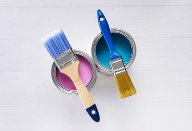 Cans of pink and light blue paints with brushes on white wooden table, flat lay