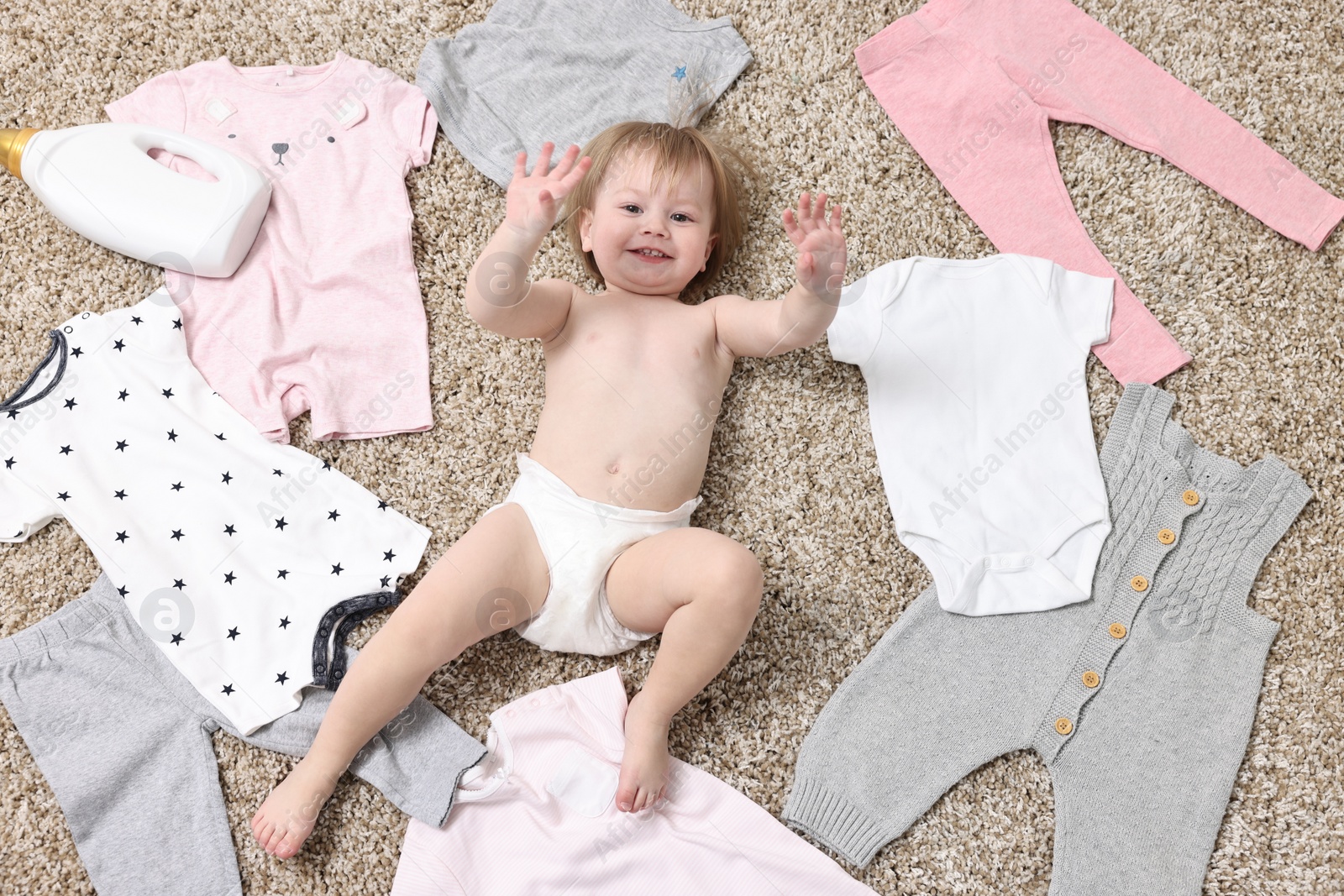 Photo of Little girl among baby clothes and detergents on carpet, top view