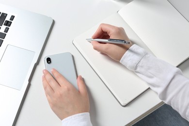 Photo of Woman with smartphone writing in notebook at white table, closeup