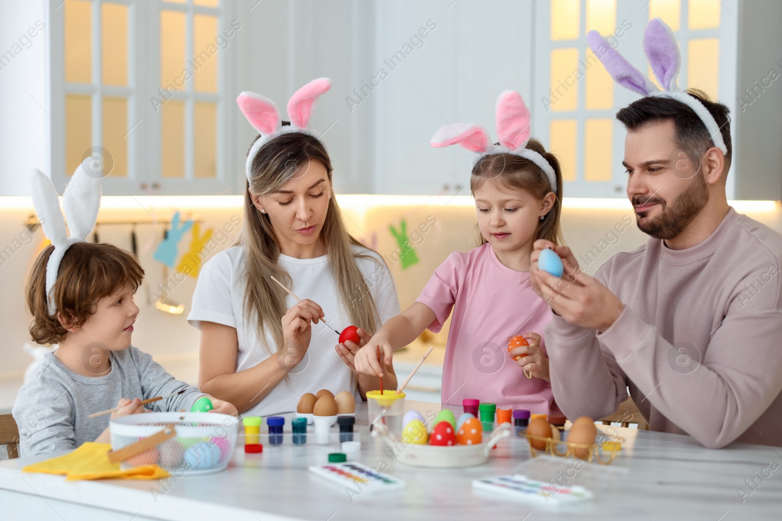 Photo of Easter celebration. Happy family with bunny ears painting eggs at white marble table in kitchen