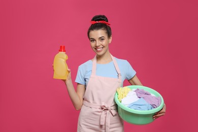 Photo of Housewife holding bottle of cleaning product and basin with clothes on pink background