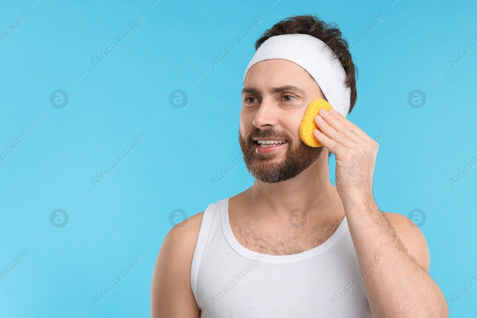 Photo of Man with headband washing his face using sponge on light blue background, space for text