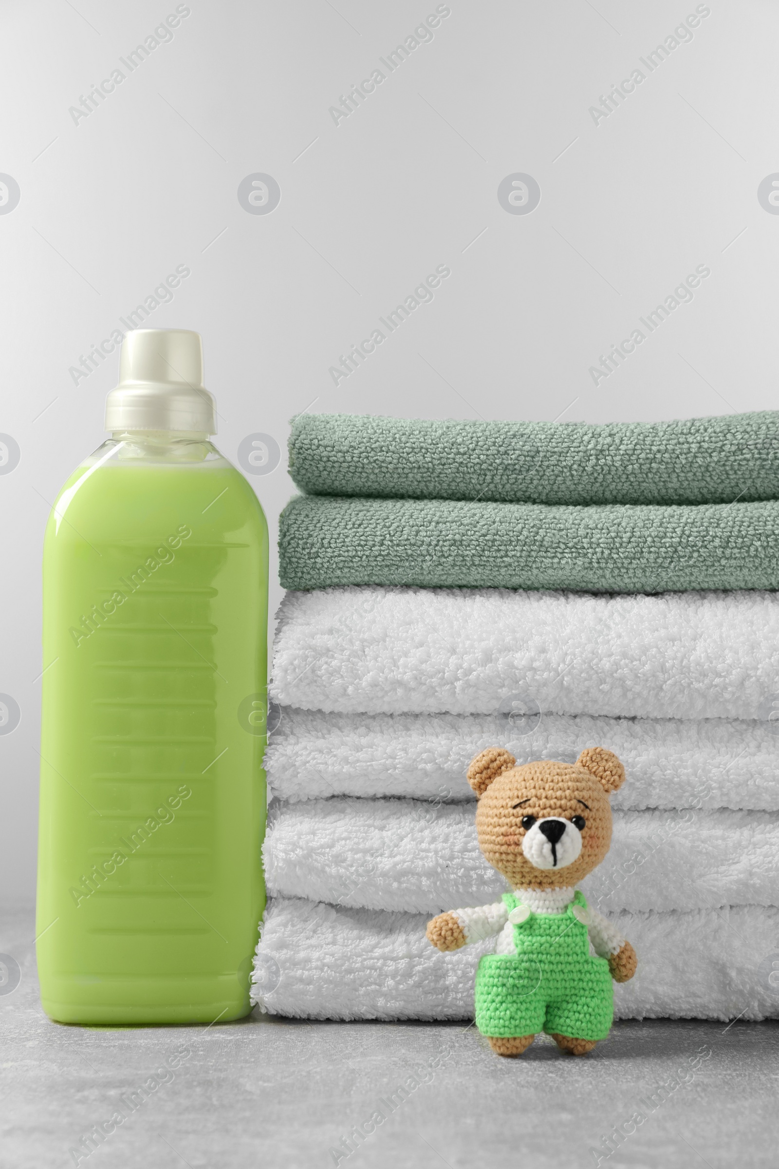 Photo of Bottle of laundry detergent, stacked fresh towels and knitted bear toy on grey table