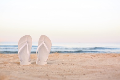 White flip flops on sand near sea, space for text. Beach accessories