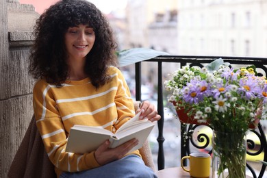 Photo of Young woman reading book at table with beautiful flowers on balcony