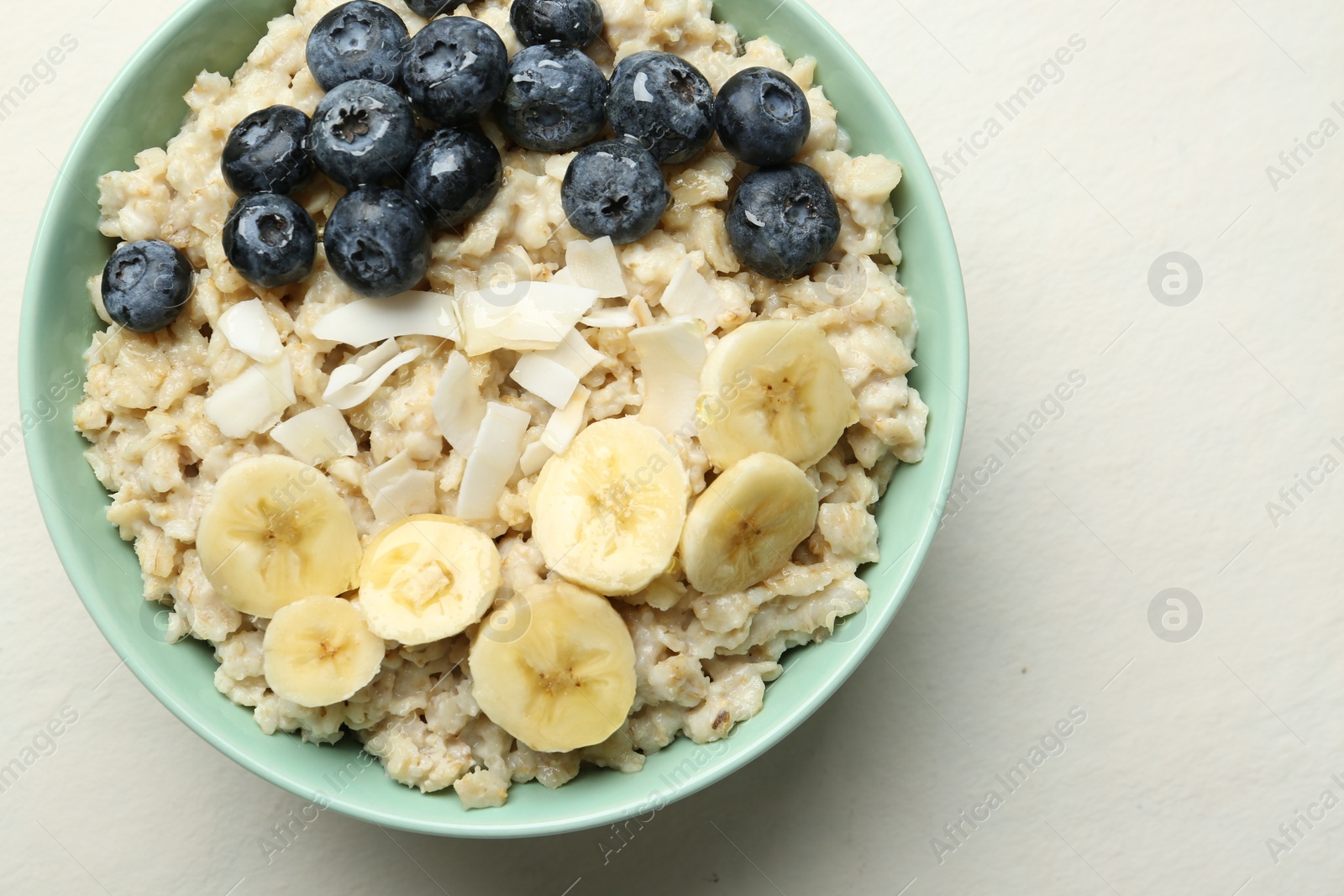 Photo of Tasty oatmeal with banana, blueberries, coconut flakes and honey served in bowl on beige table, top view. Space for text