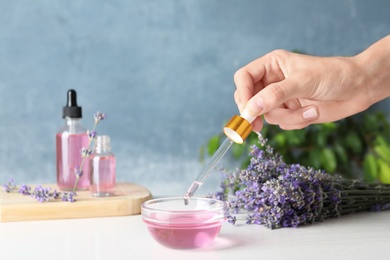 Woman holding dropper with lavender essential oil over bowl on white wooden table against blue background, closeup. Space for text