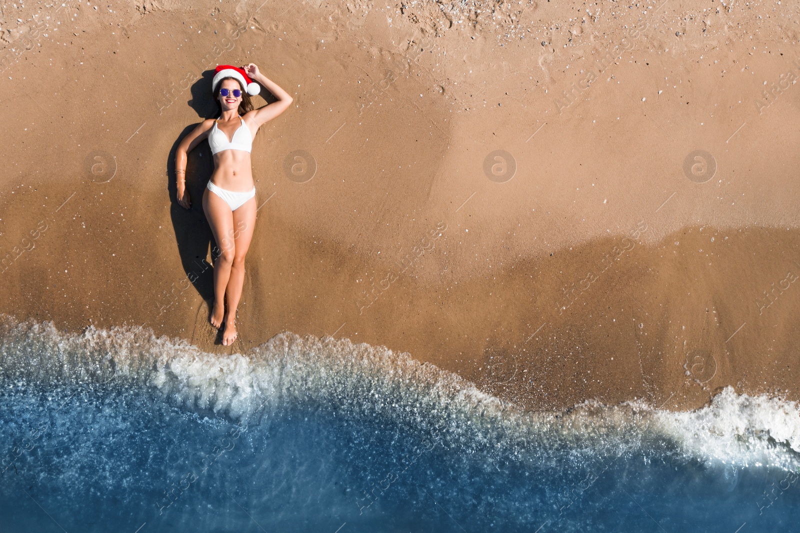 Image of Young woman wearing Santa hat and bikini on beach, top view. Christmas vacation