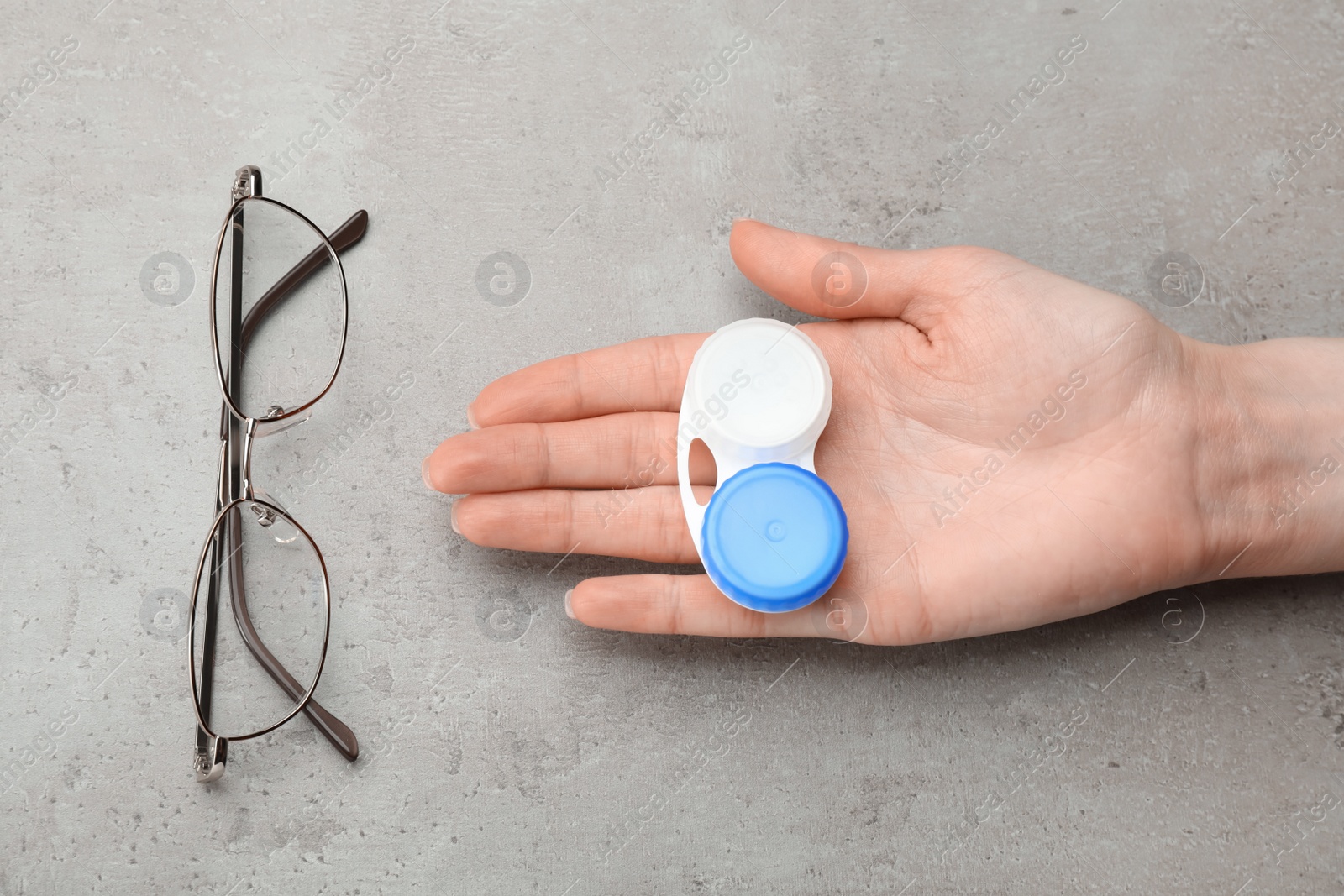 Photo of Woman holding case with contact lenses near glasses at grey table, top view