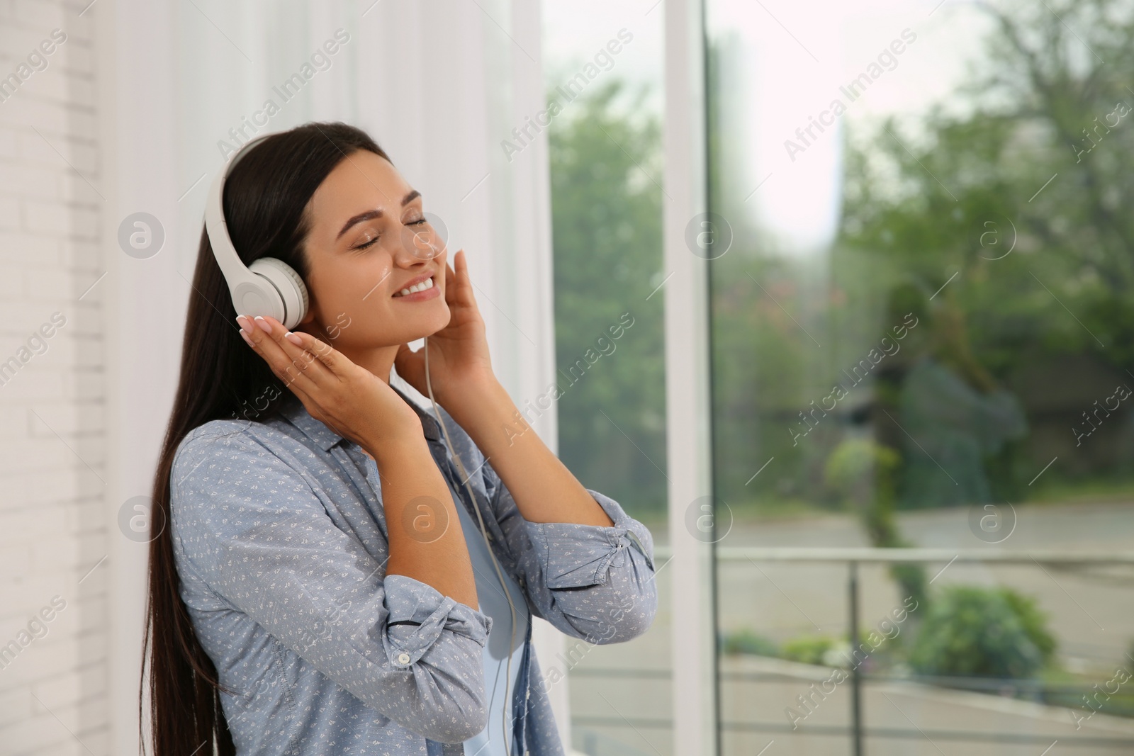 Photo of Young woman listening to music near window at home, space for text