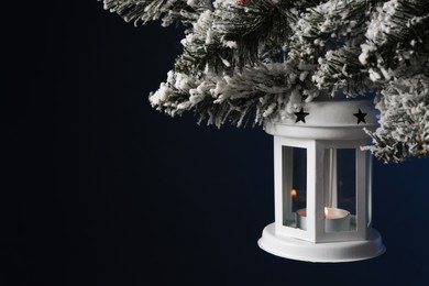 Photo of Christmas lantern with burning candle on fir tree against dark background, closeup. Space for text