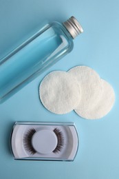 Bottle of makeup remover, cotton pads and false eyelashes on light blue background, flat lay