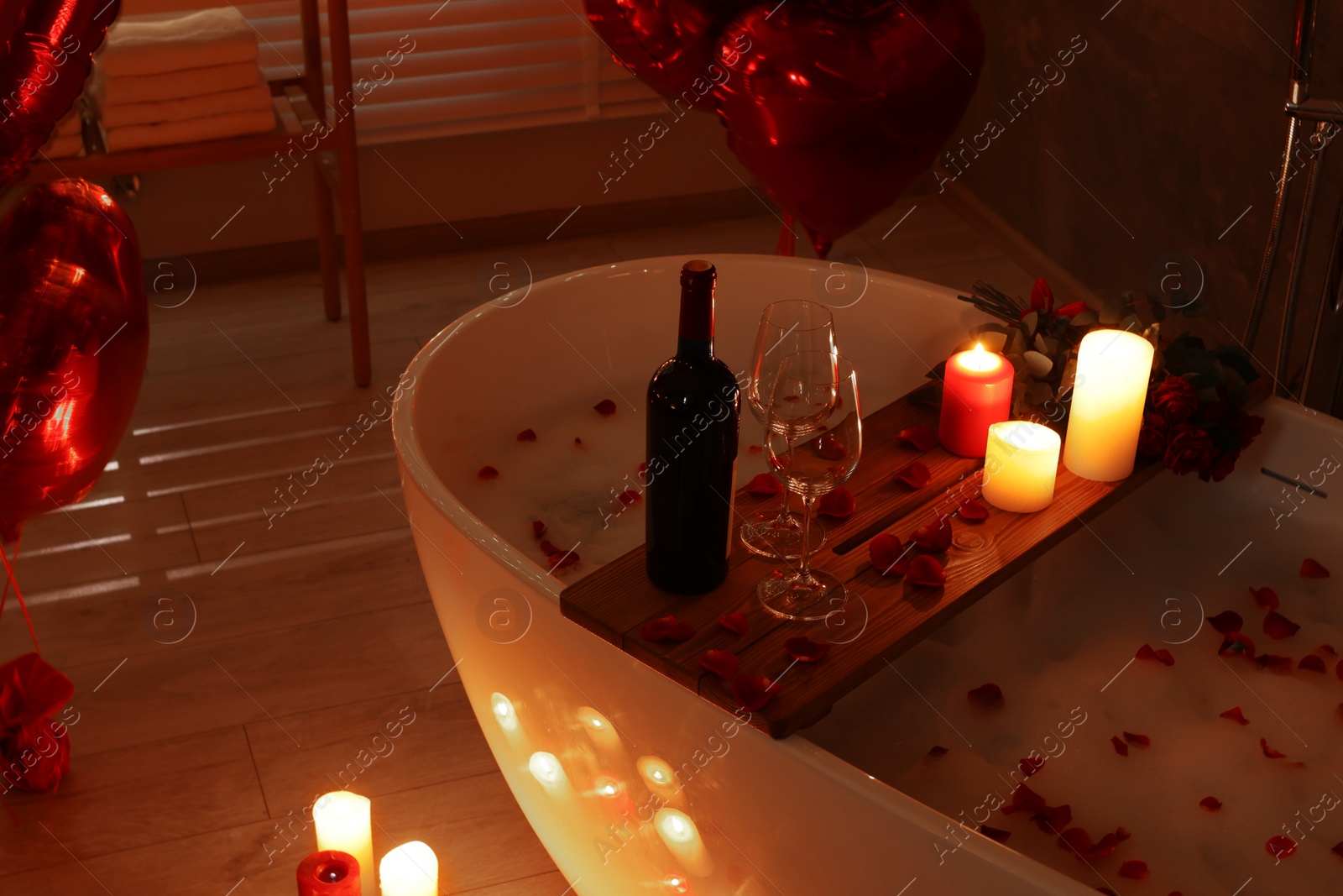 Photo of Wooden tray with wine, burning candles and rose petals on tub in bathroom. Valentine's day celebration