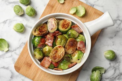 Photo of Delicious Brussels sprouts with bacon in dish on white marble table, flat lay