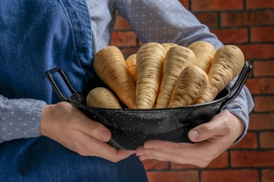 Photo of Woman holding bowl with fresh ripe parsnips near red brick wall, closeup