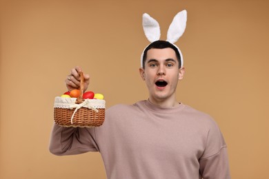 Photo of Easter celebration. Handsome young man with bunny ears holding basket of painted eggs on beige background