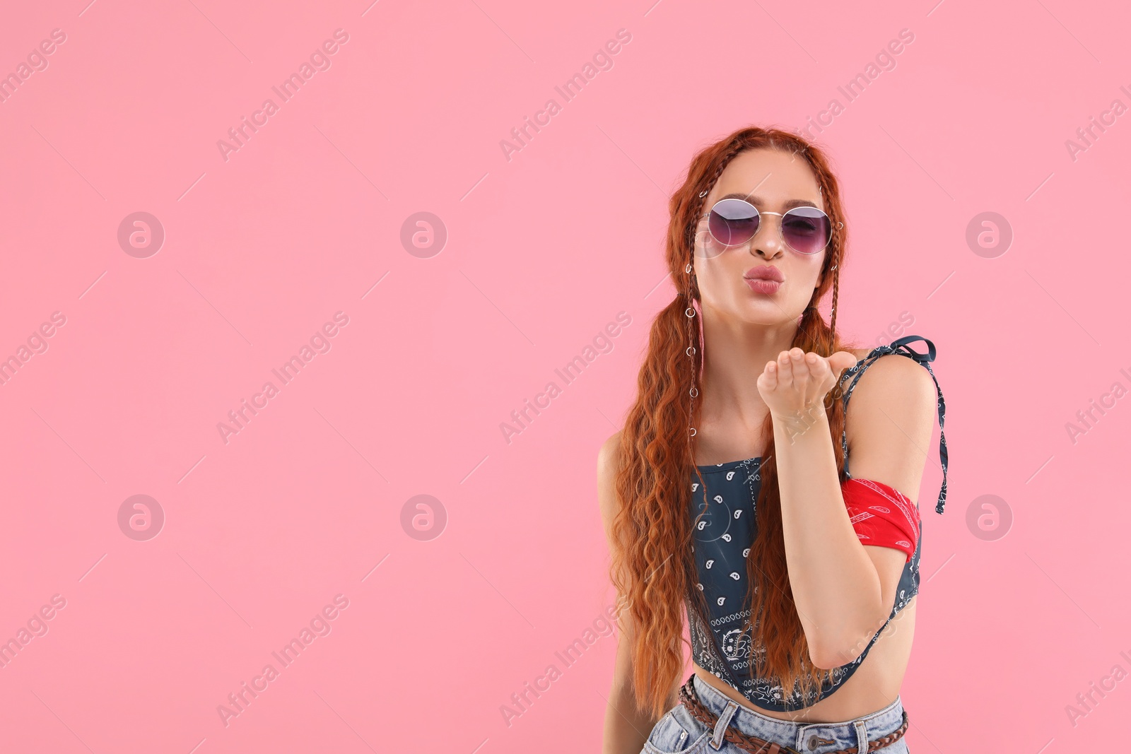 Photo of Stylish young hippie woman in sunglasses blowing kiss on pink background, space for text