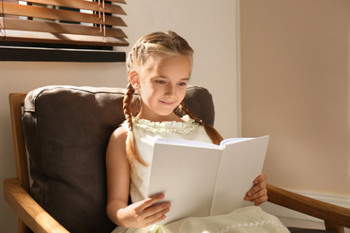 Photo of Little girl reading book in armchair at home