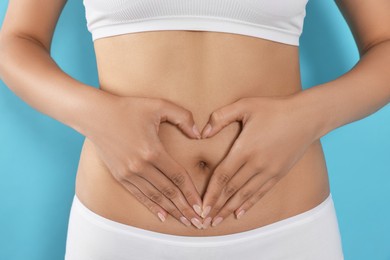 Woman in underwear making heart with hands on her belly against light blue background, closeup. Healthy stomach