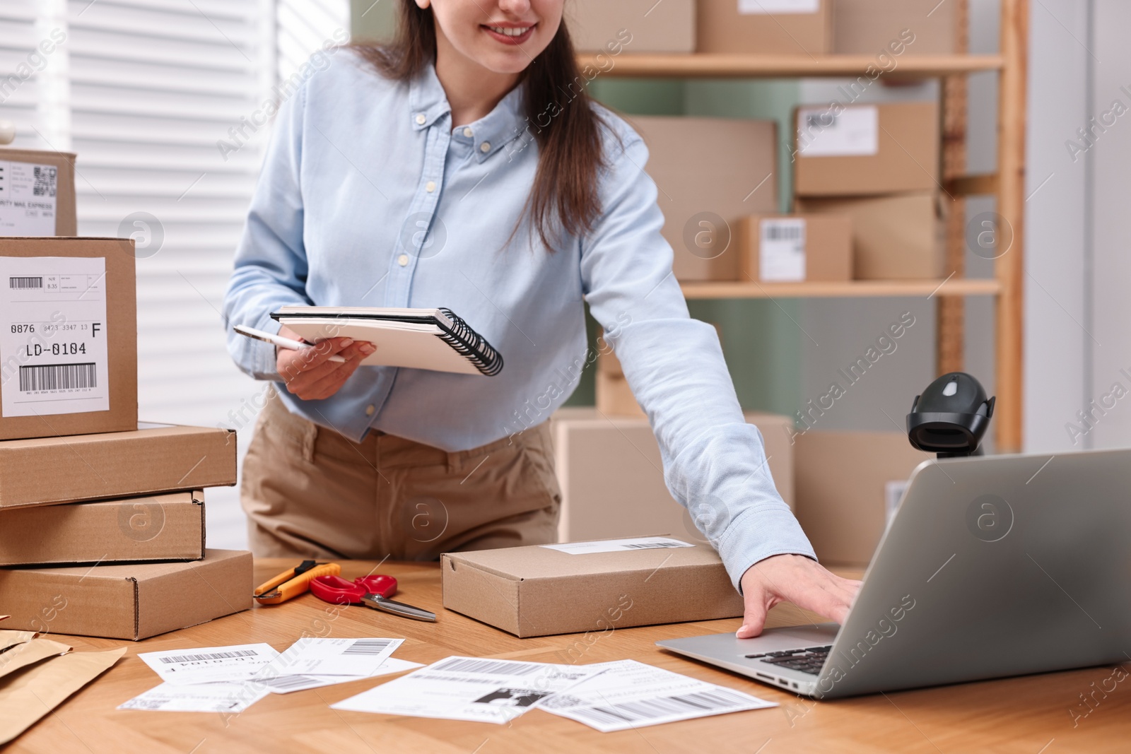 Photo of Parcel packing. Post office worker with notebook using laptop at wooden table indoors, closeup