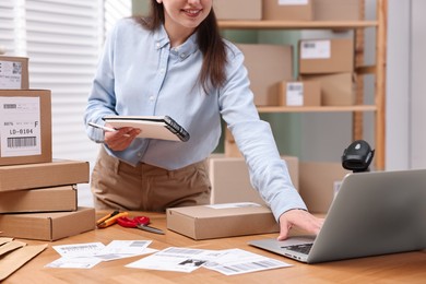 Parcel packing. Post office worker with notebook using laptop at wooden table indoors, closeup