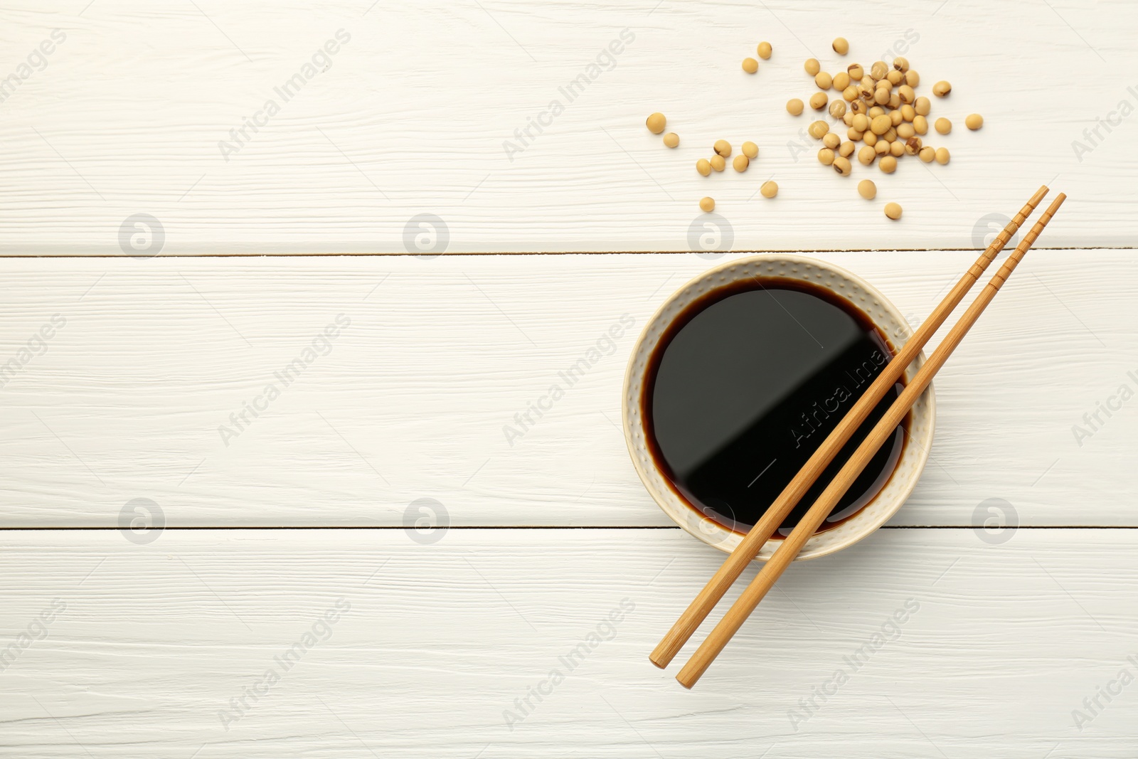 Photo of Tasty soy sauce in bowl, chopsticks and soybeans on white wooden table, top view. Space for text