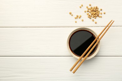 Tasty soy sauce in bowl, chopsticks and soybeans on white wooden table, top view. Space for text