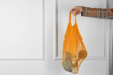 Woman holding mesh bag with different items near white wall, closeup and space for text. Conscious consumption