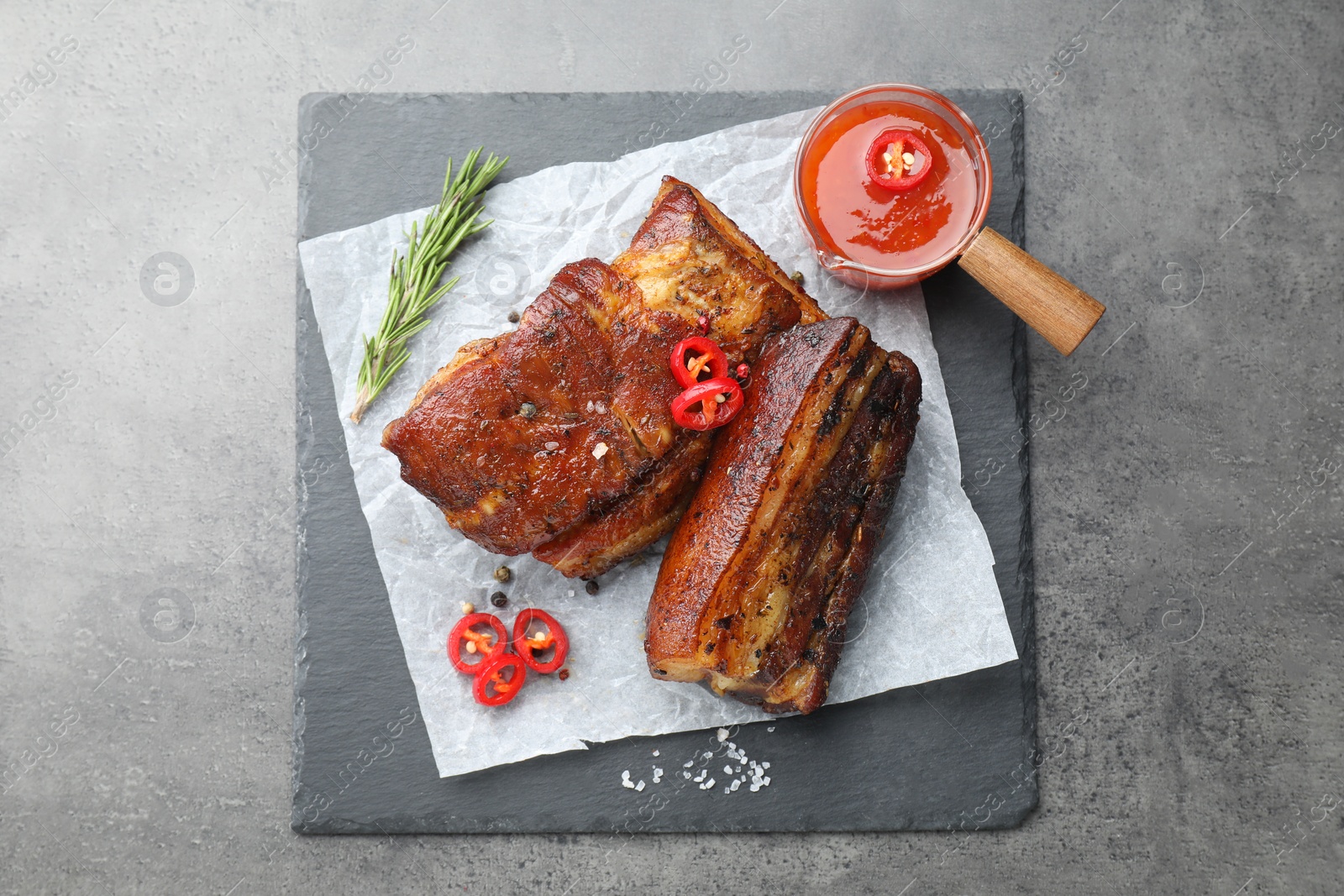 Photo of Pieces of baked pork belly served with sauce, rosemary and chili pepper on grey table, top view