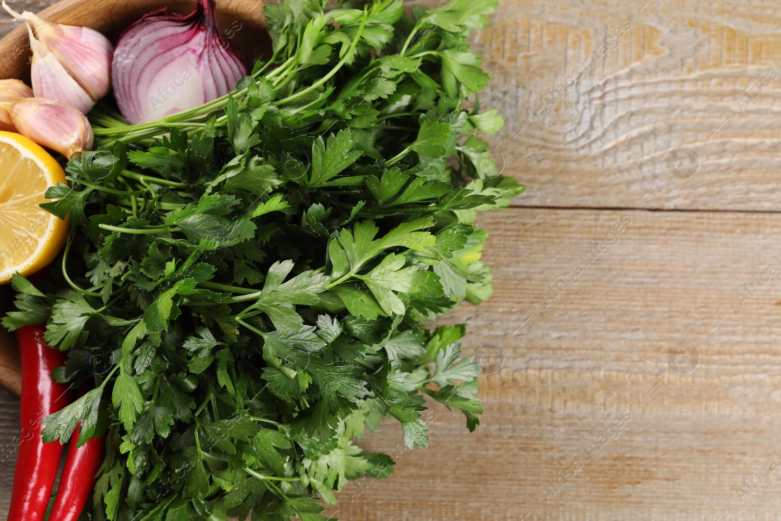 Photo of Bowl with fresh green parsley, chili peppers, lemon, onion and garlic on wooden table, top view. Space for text