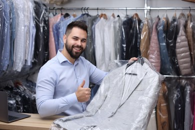 Dry-cleaning service. Happy worker holding hanger with jacket in plastic bag and showing thumb up at counter indoors