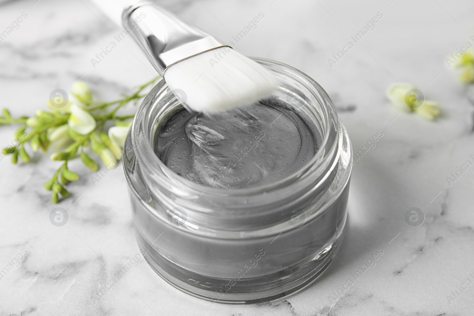 Photo of Professional face mask with brush and flowers on white marble table, closeup