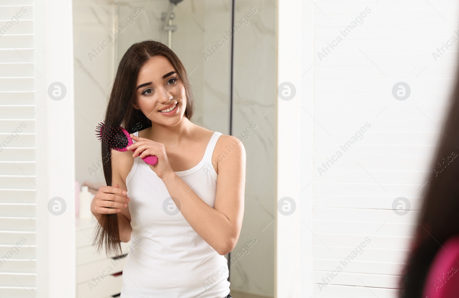 Photo of Beautiful young woman with hair brush looking into mirror in bathroom