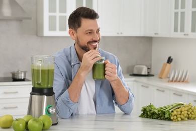 Photo of Happy man drinking delicious smoothie at white marble table in kitchen