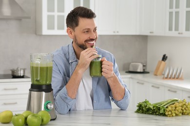 Happy man drinking delicious smoothie at white marble table in kitchen