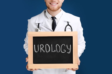 Photo of Male doctor holding small blackboard with word UROLOGY on blue background, closeup